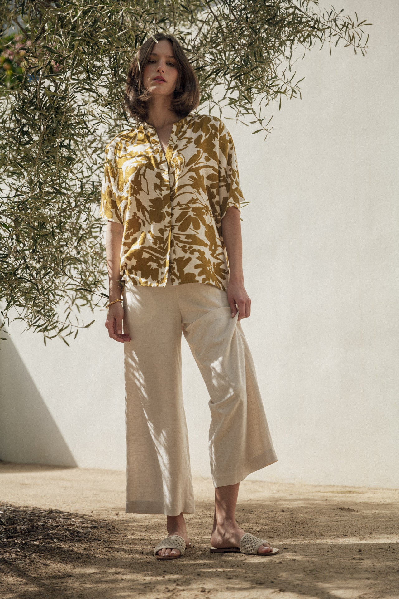 Woman wearing a yellow floral blouse and beige pants standing under olive trees.